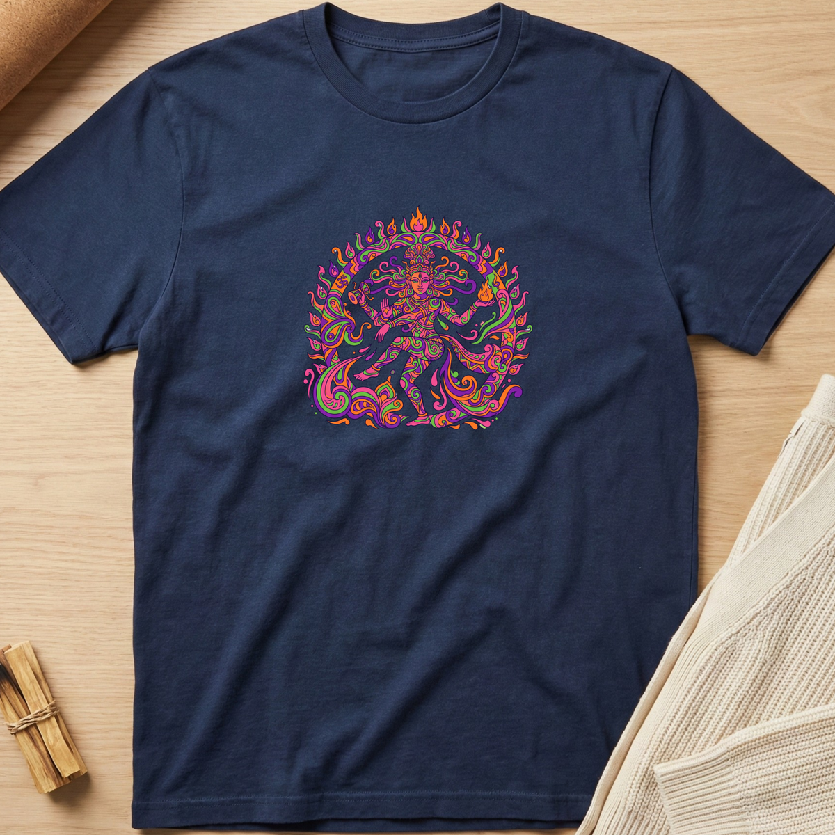 A navy blue short-sleeve t-shirt with a colorful, psychedelic-style graphic of a dancing deity in the center, laid flat on a light wood background with neutral props. Minimalist yoga apparel by pemuyo.