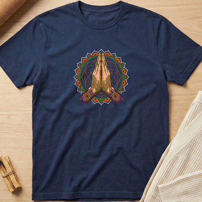 A navy blue short-sleeve t-shirt laid flat on a light wood surface, featuring a colorful graphic of two hands in a prayer pose inside a circular mandala design. Styled flat-lay props include a rolled tan material in the top-left corner and a folded cream knit fabric in the bottom-right corner. Minimalist yoga apparel by pemuyo.