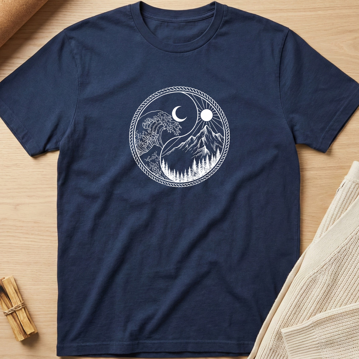 A navy blue short-sleeve t-shirt with a white circular graphic of a yin-yang style mountain and moon design, laid flat on a light wood background with styled props. Minimalist yoga apparel by pemuyo.
