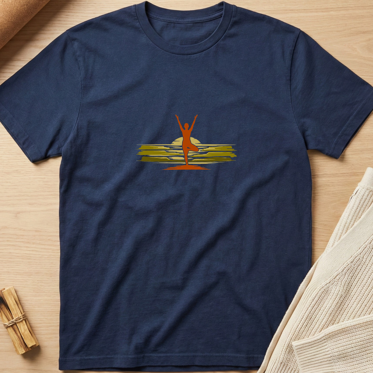 A navy blue short-sleeve crewneck t-shirt laid flat on a light wood surface, featuring a central graphic of a red silhouette of a person standing on yellow-green waves with a yellow sun behind them. Minimalist yoga apparel by pemuyo.