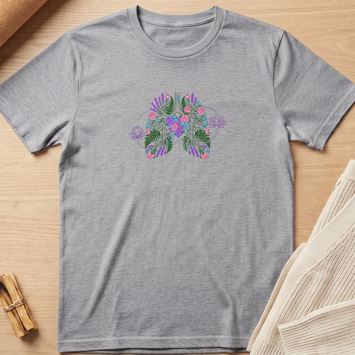 A heather gray short-sleeve crewneck t-shirt featuring a colorful butterfly graphic on the chest with green leaves, pink flowers, and purple accents, laid flat on a light wood background. Minimalist yoga apparel by pemuyo.