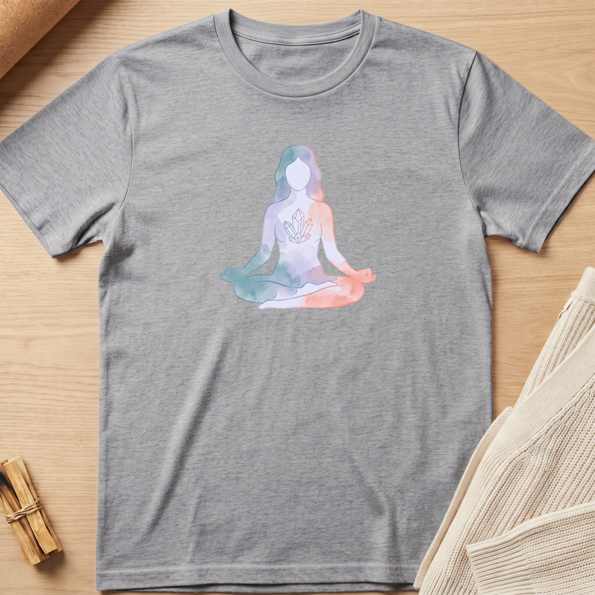 A heather gray short-sleeve t-shirt laid flat on a light wood surface, featuring a pastel watercolor-style graphic of a seated meditating figure on the chest. The shirt is styled with neutral props, including a rolled fabric corner and a small bundle of dried stems. Minimalist yoga apparel by pemuyo.