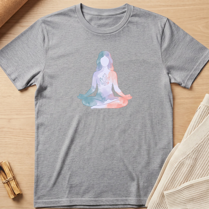 A heather gray short-sleeve t-shirt laid flat on a light wood surface, featuring a pastel watercolor-style graphic of a seated meditating figure on the chest. The shirt is styled with neutral props, including a rolled fabric corner and a small bundle of dried stems. Minimalist yoga apparel by pemuyo.