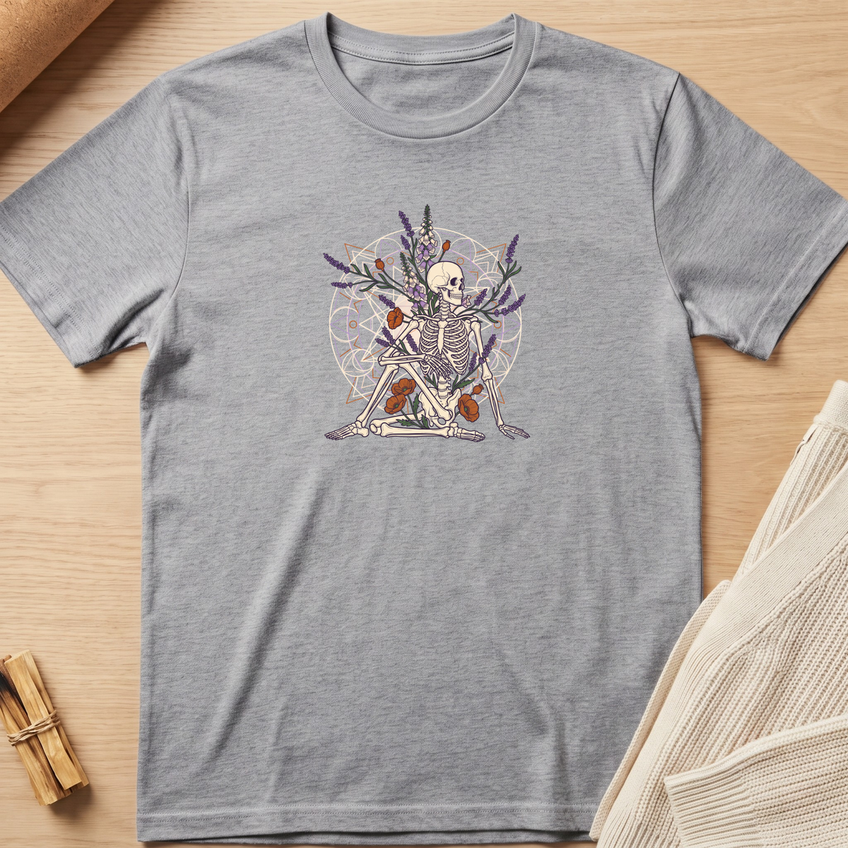 A heather gray short-sleeve t-shirt with a central graphic of a seated skeleton surrounded by lavender, orange flowers, and a circular mandala pattern, laid flat on a light wood background with styled props. Minimalist yoga apparel by pemuyo.