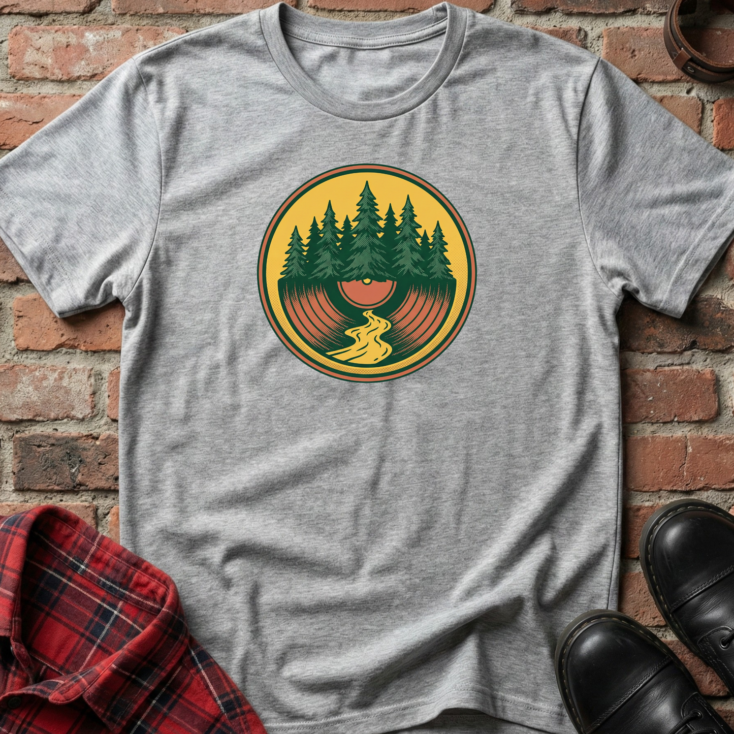 Gray t-shirt with a graphic design of trees and a person on a vinyl record against a brick wall. Minimalist music apparel by pemuyo.