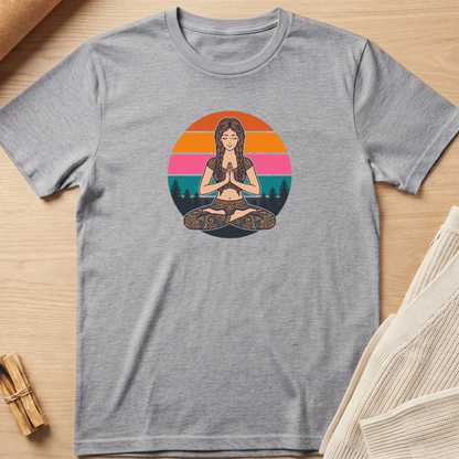 A heather gray short-sleeve t-shirt laid flat on a light wood surface, featuring a colorful graphic of a woman meditating in front of a sunset circle. The shirt is styled with a small bundle of dried herbs on the left and a folded cream knit fabric on the right. Minimalist yoga apparel by pemuyo.