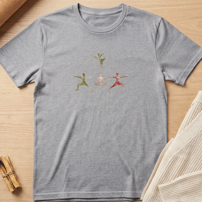A heather gray short-sleeve crewneck t-shirt featuring a graphic of three watercolor-style dancers in green, pink, and orange, laid flat on a light wood background with styled props. Minimalist yoga apparel by pemuyo.