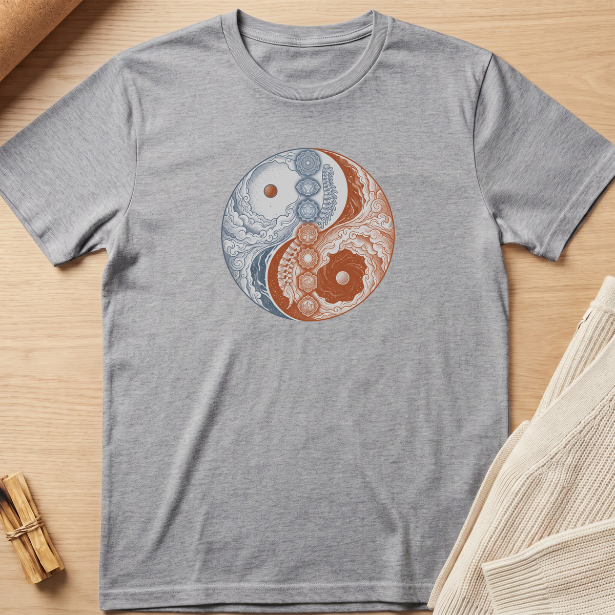 A heather gray short-sleeve t-shirt laid flat on a light wood surface, featuring a circular yin-yang style graphic in the center with blue and rust-orange ornamental details. Minimalist yoga apparel by pemuyo.