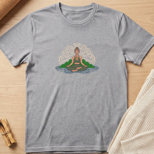 A heather gray short-sleeve t-shirt with a central graphic of a meditating figure surrounded by green hills and blue water, laid flat on a light wood surface with neutral props. Minimalist yoga apparel by pemuyo.