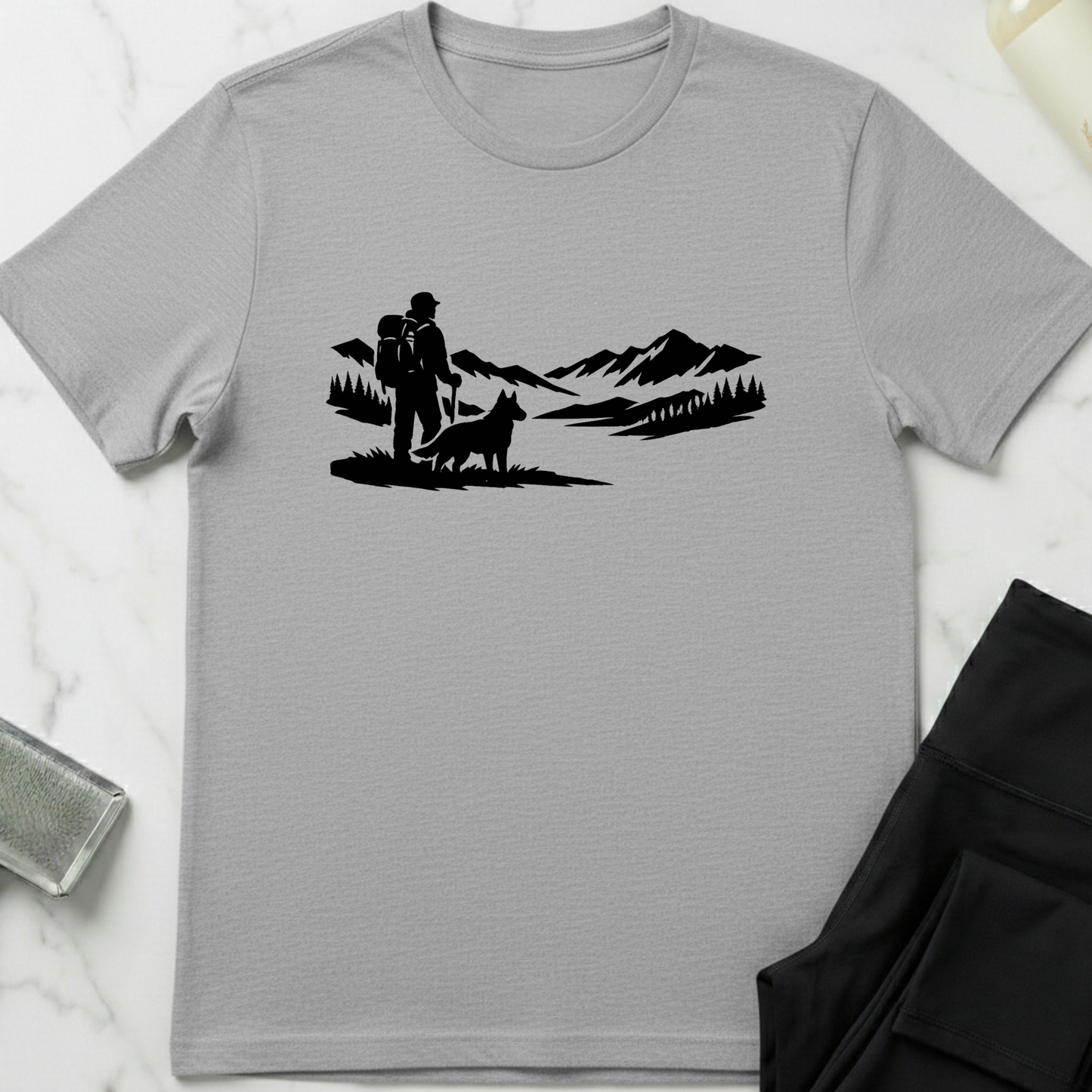 Gray t-shirt with black graphic of a couple and dog in a mountainous landscape on a white marble surface. Minimalist dog apparel by pemuyo.