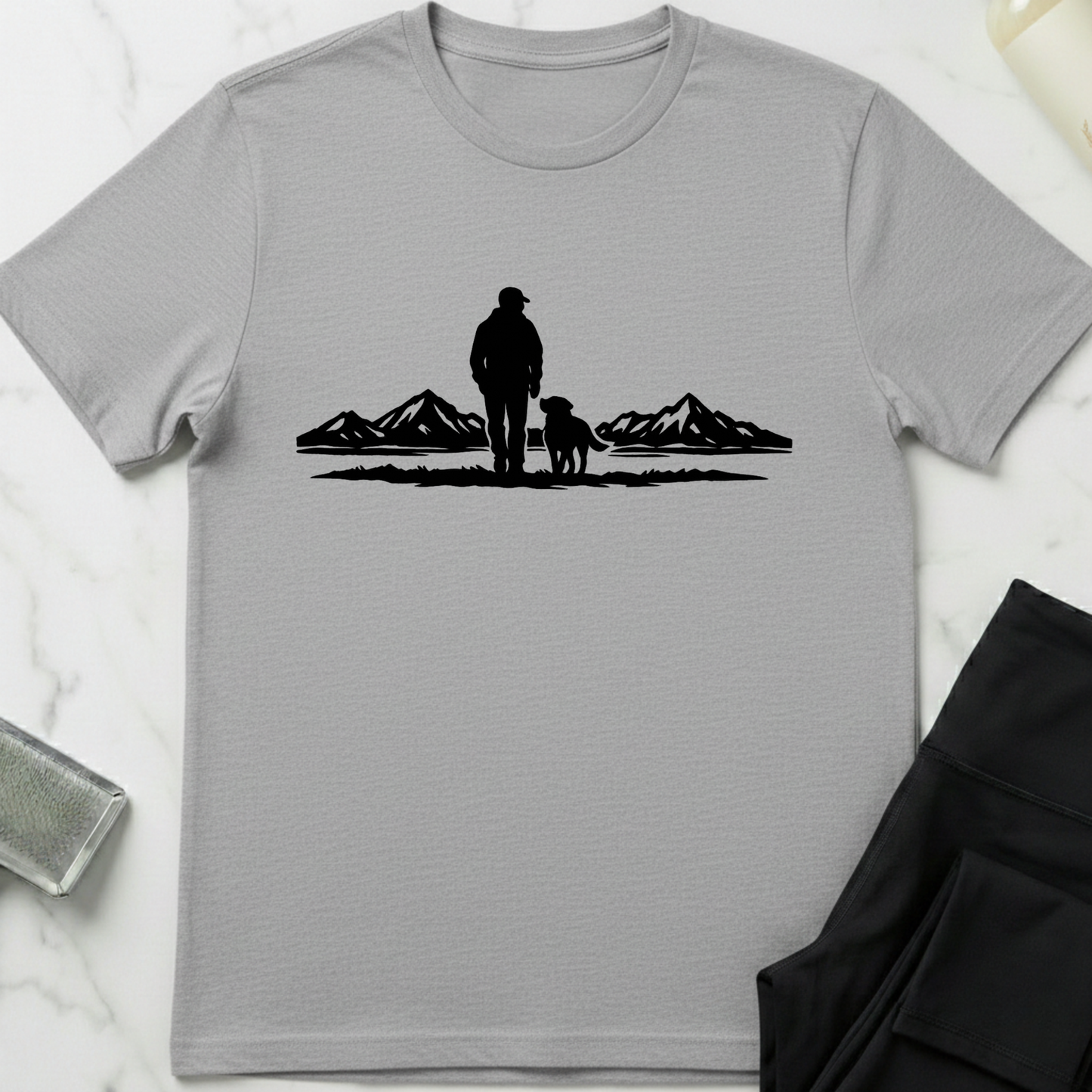 Gray t-shirt with a black silhouette of a person and dog standing in front of mountains on a white marble surface. Minimalist dog apparel by pemuyo.