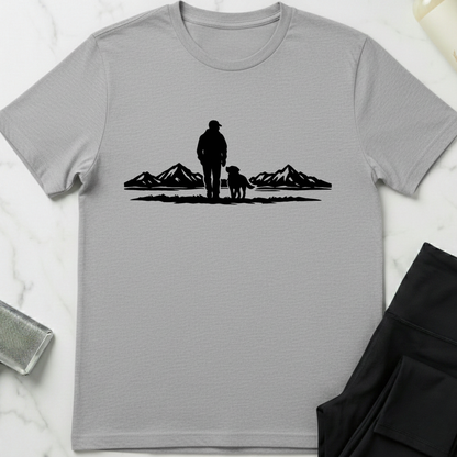Gray t-shirt with a black silhouette of a person and dog standing in front of mountains on a white marble surface. Minimalist dog apparel by pemuyo.