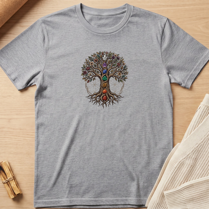 A heather gray short-sleeve t-shirt with a colorful 'Tree of Life' graphic featuring gemstone-like colors on the chest, laid flat on a light wood background with neutral props. Minimalist yoga apparel by pemuyo.