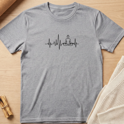 A heather gray short-sleeve t-shirt with a black line art graphic of a meditating figure on an EKG heartbeat line, displayed flat on a light wood background with neutral props. Minimalist yoga apparel by pemuyo.