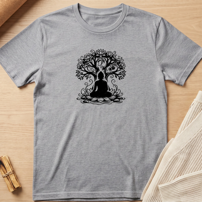 A heather gray short-sleeve t-shirt with a black graphic of a meditating figure under a tree, laid flat on a light wood background with neutral props. Minimalist yoga apparel by pemuyo.