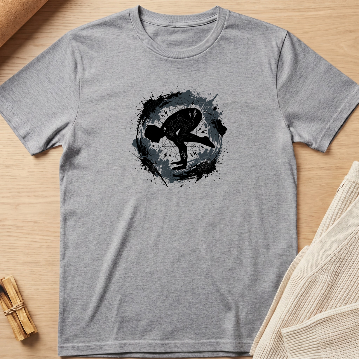 A heather gray short-sleeve t-shirt with a black and dark gray circular graphic print on the chest, laid flat on a light wood background with styled props. Minimalist yoga apparel by pemuyo.