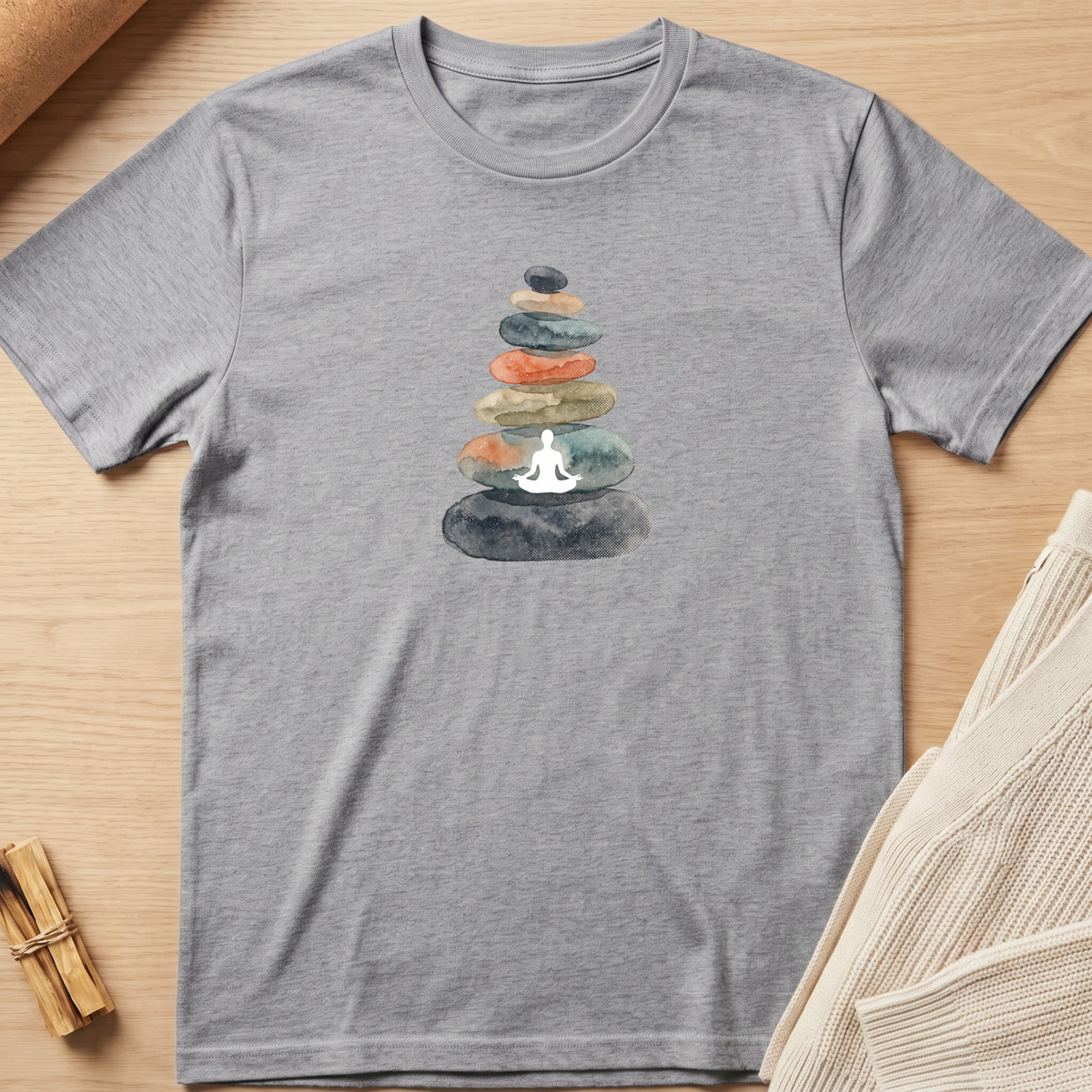 A heather gray short-sleeve t-shirt laid flat on a light wood surface, featuring a watercolor graphic of stacked stones with a white meditating figure in the center. A small bundle of dried herbs and a folded cream knit fabric are partially visible as props. Minimalist yoga apparel by pemuyo.