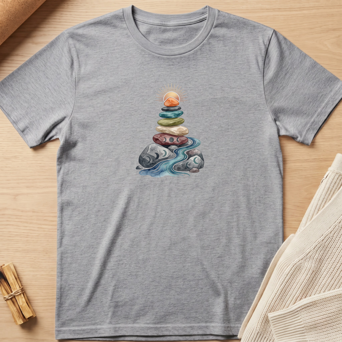 A heather gray short-sleeve t-shirt laid flat on a light wood surface, featuring a central graphic of stacked colorful stones with a glowing sun and blue water flowing beneath them. Minimalist yoga apparel by pemuyo.
