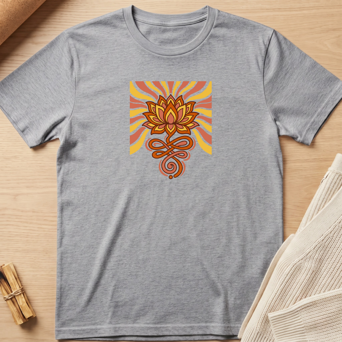 A heather gray short-sleeve t-shirt laid flat on a light wood surface, featuring a central graphic of a stylized orange and brown lotus flower with yellow and orange sunburst rays. Minimalist yoga apparel by pemuyo.