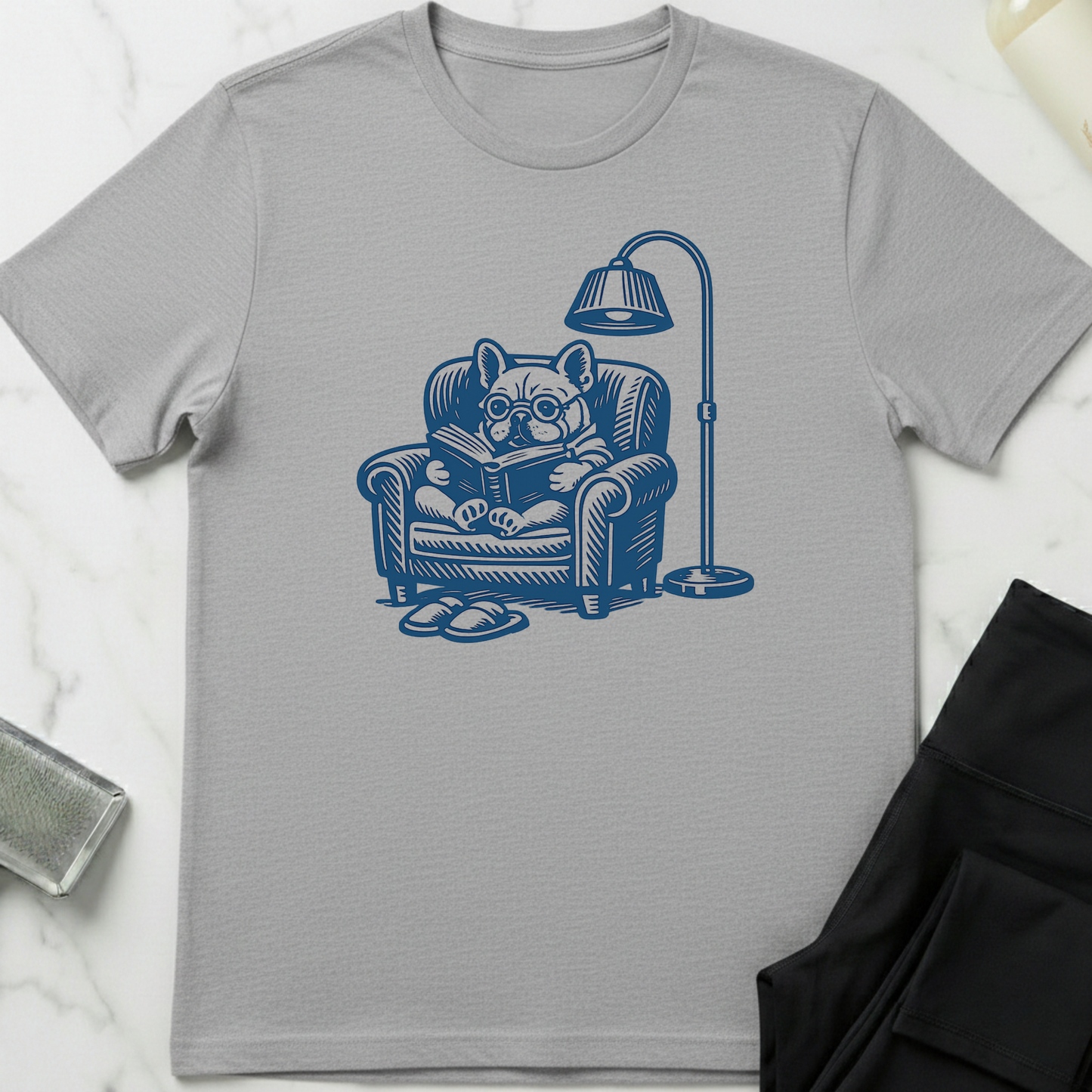 Gray t-shirt with a blue graphic of a dog reading a book on a chair, placed on a white surface. Minimalist dog apparel by pemuyo.