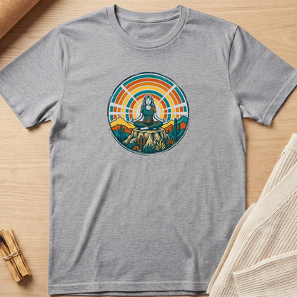 A heather gray short-sleeve t-shirt laid flat on a light wood surface, featuring a colorful circular graphic of a meditating woman surrounded by mountains and a sunset. Minimalist yoga apparel by pemuyo.