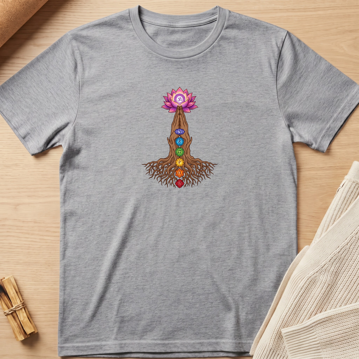 A heather gray short-sleeve t-shirt laid flat on a light wood surface, featuring a central graphic of a pink lotus flower above a brown tree trunk with colorful chakra stones and roots. A small bundle of dried herbs and a folded cream knit fabric are partially visible at the edges. Minimalist yoga apparel by pemuyo.