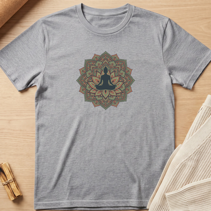 A heather gray short-sleeve t-shirt with a central mandala graphic featuring a meditating silhouette in dark blue, surrounded by green, orange, and beige details. The t-shirt is laid flat on a light wood surface with a small bundle of incense sticks on the left and a folded cream knit blanket on the right. Minimalist yoga apparel by pemuyo.