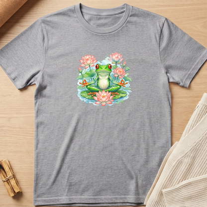 A heather gray short-sleeve t-shirt laid flat on a light wood surface, featuring a colorful graphic of a green frog meditating on a lily pad surrounded by pink water lilies. A beige knit sweater is partially visible on the right, and a small bundle of sticks is on the left. Minimalist yoga apparel by pemuyo.