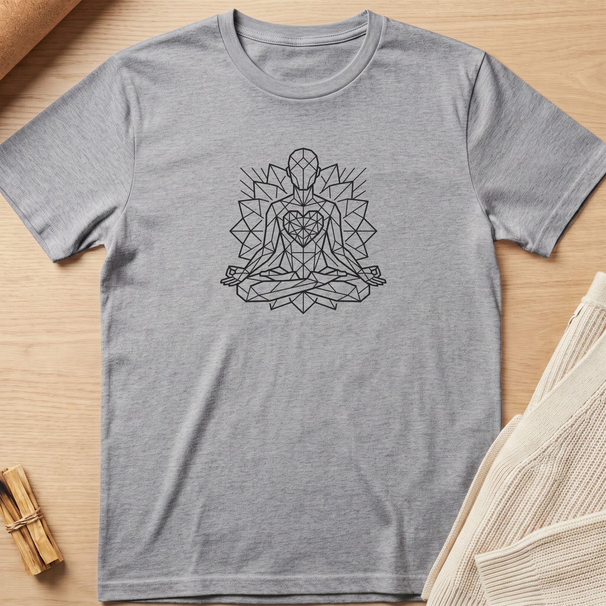 A heather gray short-sleeve t-shirt with a black geometric line art graphic of a meditating figure, laid flat on a light wood background with styled props. Minimalist yoga apparel by pemuyo.