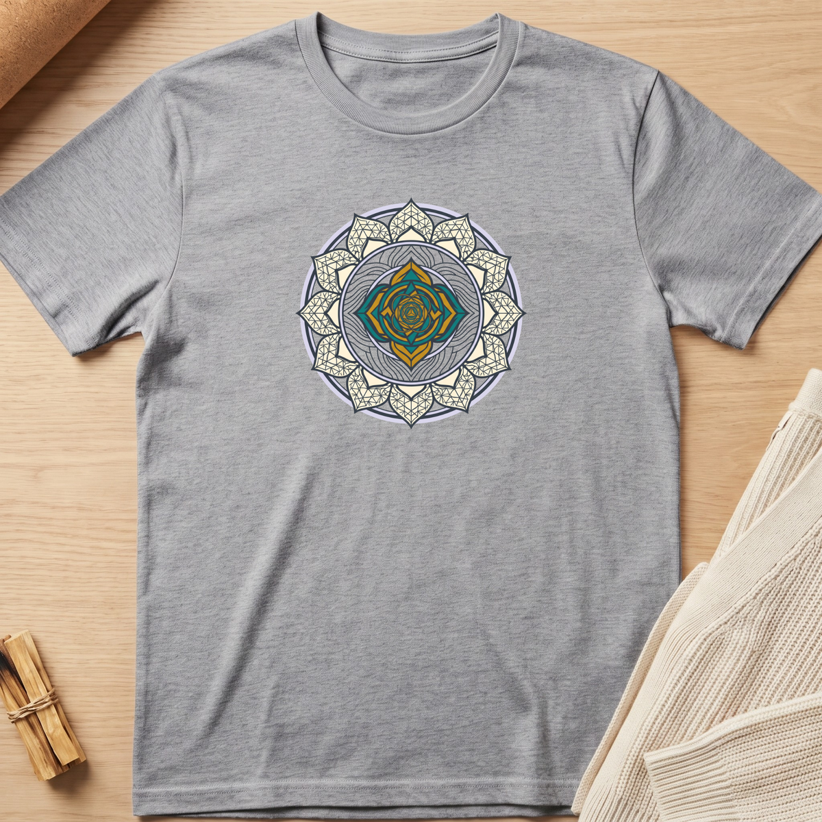 A heather gray short-sleeve t-shirt with a detailed mandala graphic in gold, teal, and white on the chest, laid flat on a light wood background with neutral props. Minimalist yoga apparel by pemuyo.