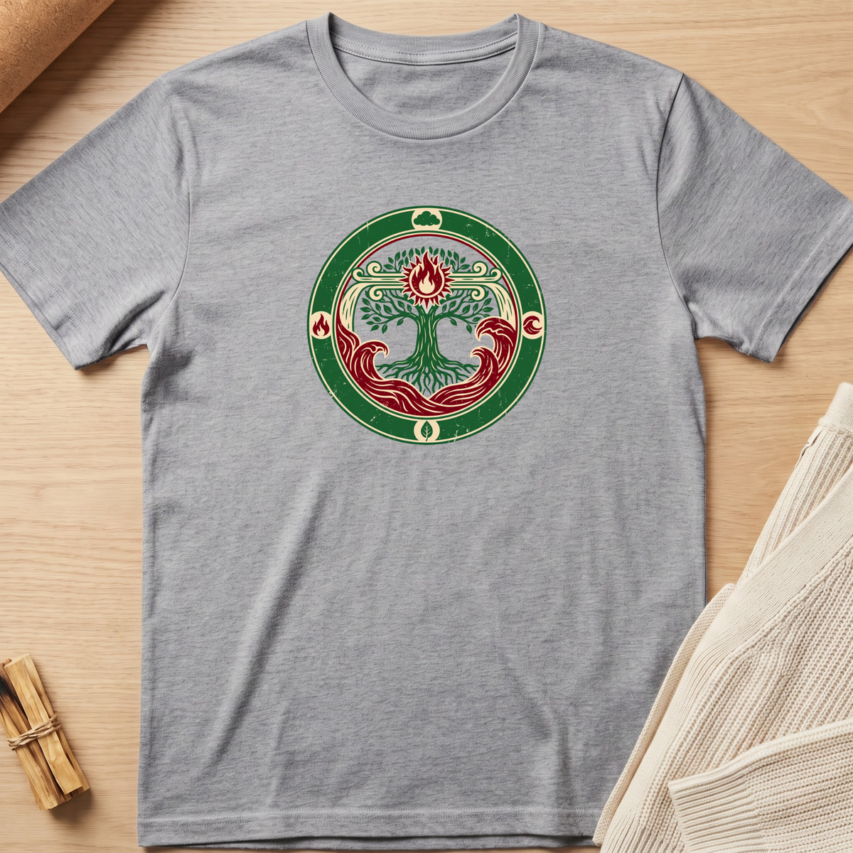 A heather gray short-sleeve t-shirt with a circular graphic on the chest featuring a stylized tree of life in green, red, and cream, laid flat on a light wood background with neutral props. Minimalist yoga apparel by pemuyo.