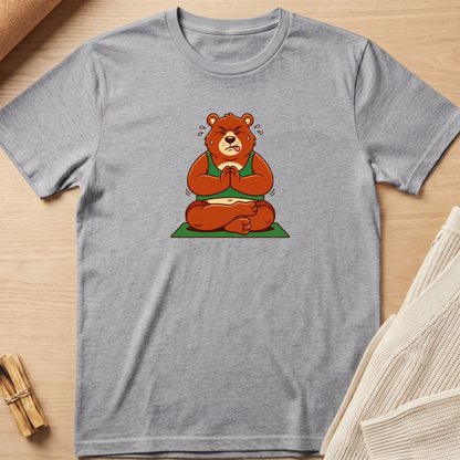 A heather gray short-sleeve t-shirt laid flat on a light wood surface, featuring a cartoon graphic of a brown bear meditating cross-legged on a green mat. Minimalist yoga apparel by pemuyo.