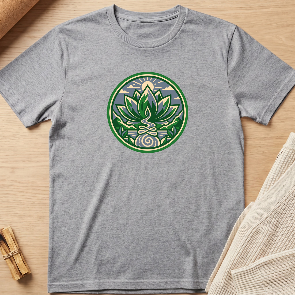 A heather gray crewneck t-shirt featuring a circular green and white lotus medallion graphic on the chest, laid flat on a light wood background with neutral props. Minimalist yoga apparel by pemuyo.