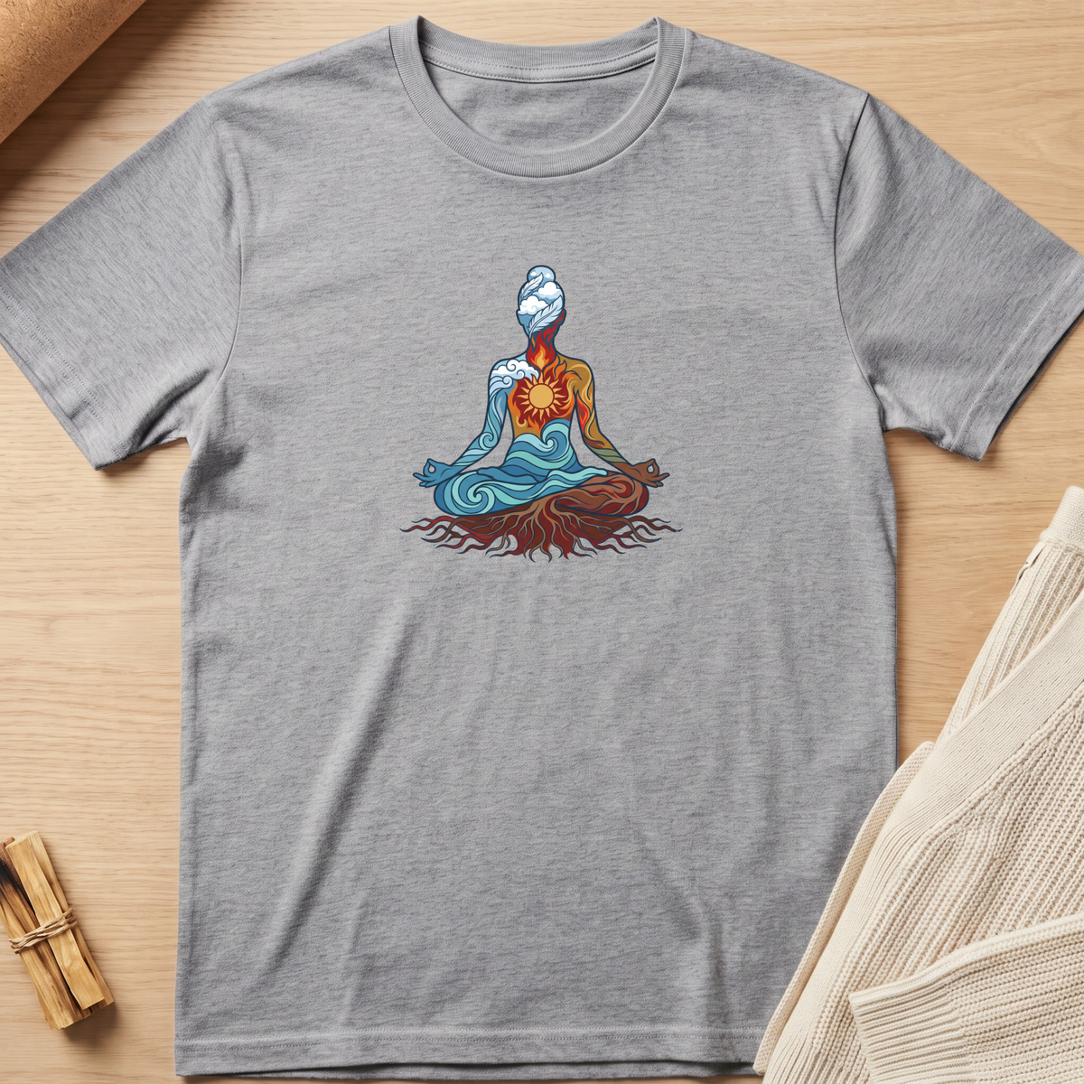 A heather gray short-sleeve t-shirt featuring a colorful graphic of a meditating figure with blue and orange tones, laid flat on a light wood background with styled props. Minimalist yoga apparel by pemuyo.