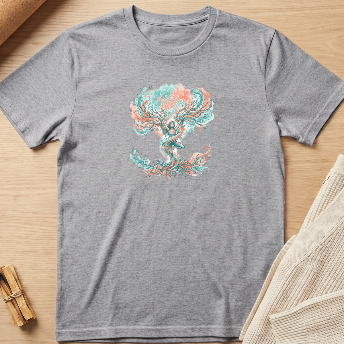 A heather gray short-sleeve t-shirt with a colorful graphic of a tree with a meditating figure inside, styled flat-lay on a light wood background with a knit blanket and a small bundle of sticks. Minimalist yoga apparel by pemuyo.
