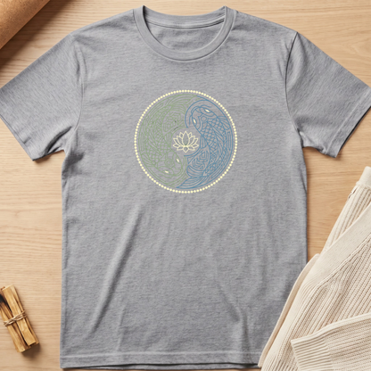 A heather gray short-sleeve t-shirt laid flat on a light wood surface, featuring a circular yin-yang style graphic with a lotus flower in the center. The left half of the circle is green with blue line details, and the right half is blue with green line details, accented by a dotted border. Minimalist yoga apparel by pemuyo.