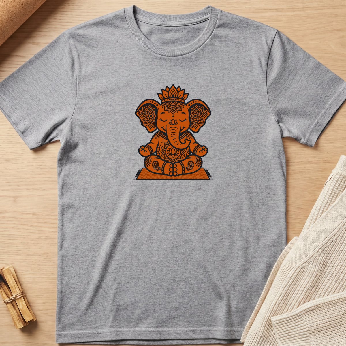 A heather gray short-sleeve t-shirt featuring a central graphic of Ganesha in orange and brown tones, styled flat on a light wood background with a small bundle of cinnamon sticks and a folded cream knit fabric visible at the edges. Minimalist yoga apparel by pemuyo.