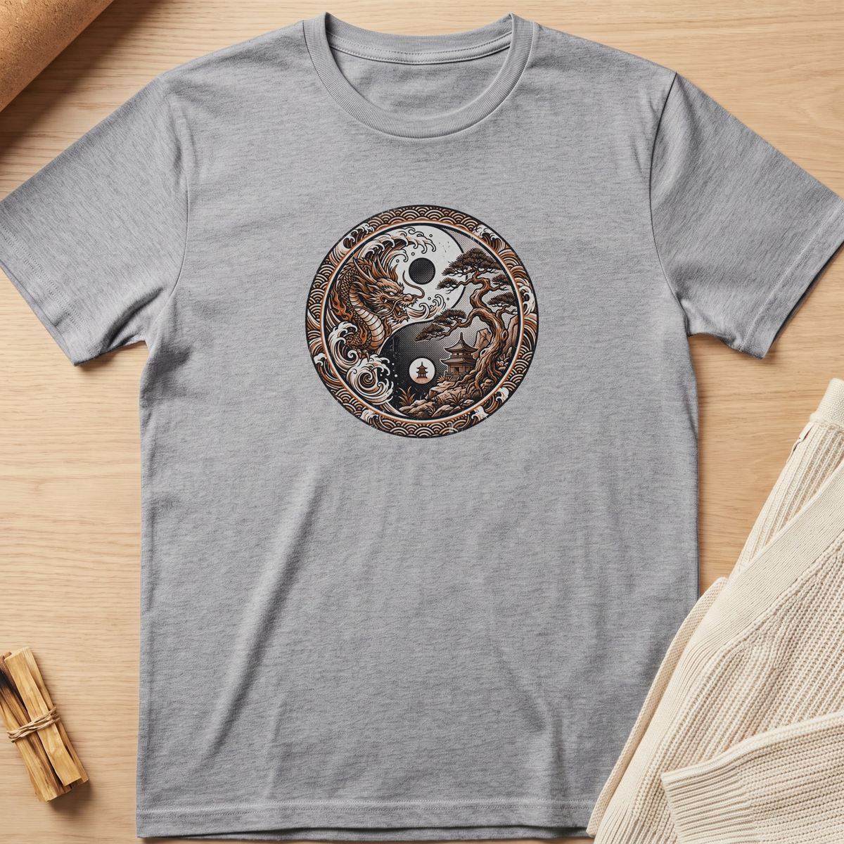 A heather gray short-sleeve t-shirt laid flat on a light wood surface, featuring a circular graphic print on the chest with a yin-yang design made from a skull, tree of life, and stars. Minimalist yoga apparel by pemuyo.