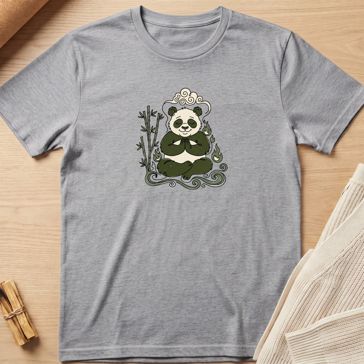 A heather gray short-sleeve t-shirt with a central graphic of a panda meditating in front of bamboo, styled flat on a light wood background with a knit sweater and cinnamon sticks. Minimalist yoga apparel by pemuyo.