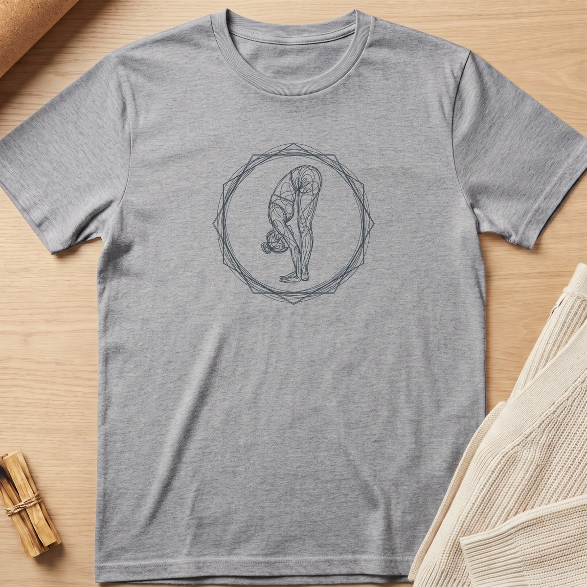 A heather gray short-sleeve t-shirt with a dark gray line art graphic of a person in a yoga forward bend pose inside a geometric circle, laid flat on a light wood background with neutral props. Minimalist yoga apparel by pemuyo.