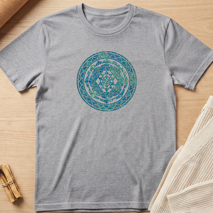 A heather gray short-sleeve t-shirt with a circular blue and green geometric mandala print on the chest, laid flat on a light wood background with styled props. Minimalist yoga apparel by pemuyo.