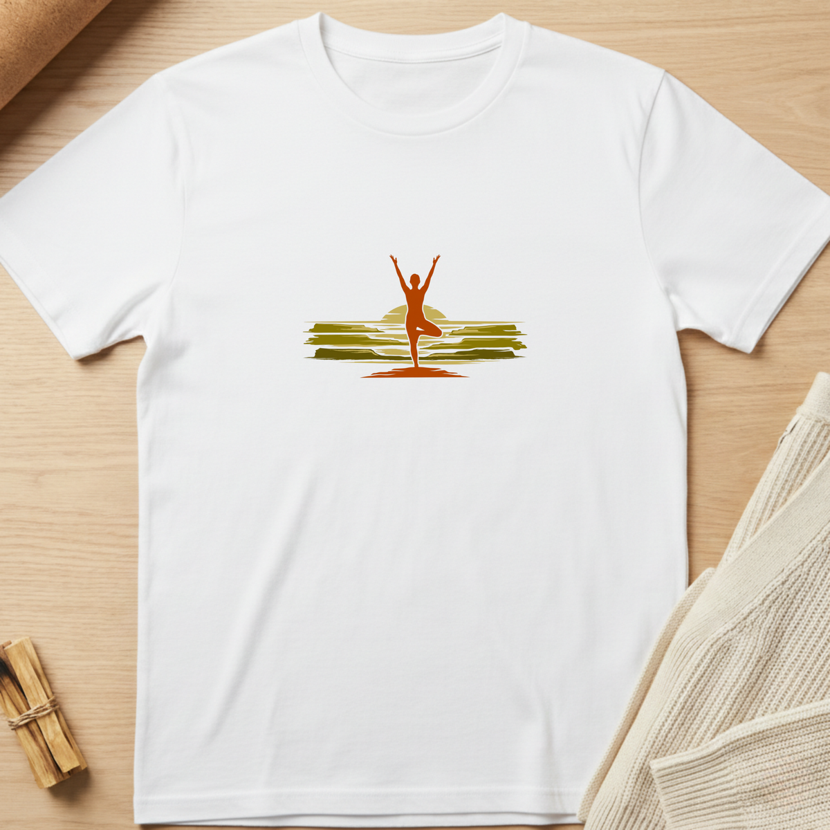 A white short-sleeve crewneck t-shirt laid flat on a light wood surface, featuring a central graphic of a yoga pose silhouette in orange-red against a yellow sun and green hills. Minimalist yoga apparel by pemuyo.