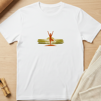 A white short-sleeve crewneck t-shirt laid flat on a light wood surface, featuring a central graphic of a yoga pose silhouette in orange-red against a yellow sun and green hills. Minimalist yoga apparel by pemuyo.