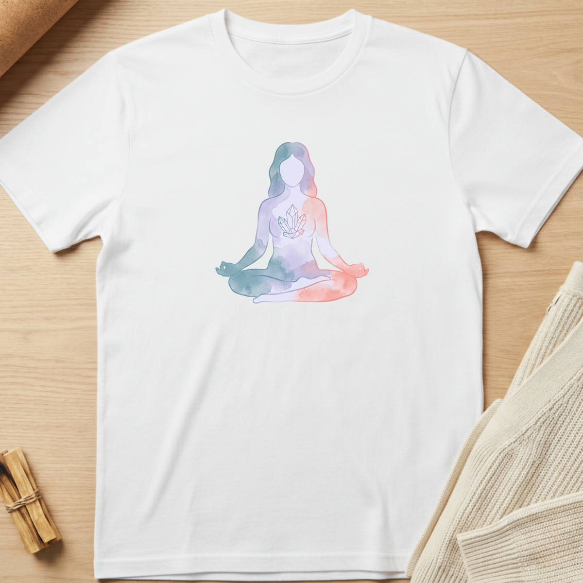 A white short-sleeve crewneck t-shirt laid flat on a light wood surface, featuring a watercolor-style graphic of a meditating figure in pastel blue, purple, and pink tones on the chest. Minimalist yoga apparel by pemuyo.