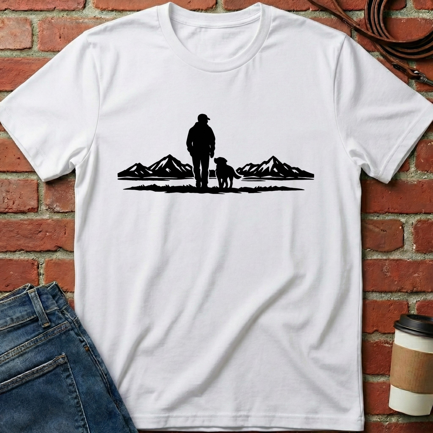 White t-shirt with a black graphic of a person and dog standing in front of mountains, placed against a brick wall. Minimalist dog apparel by pemuyo.