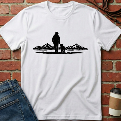 White t-shirt with a black graphic of a person and dog standing in front of mountains, placed against a brick wall. Minimalist dog apparel by pemuyo.