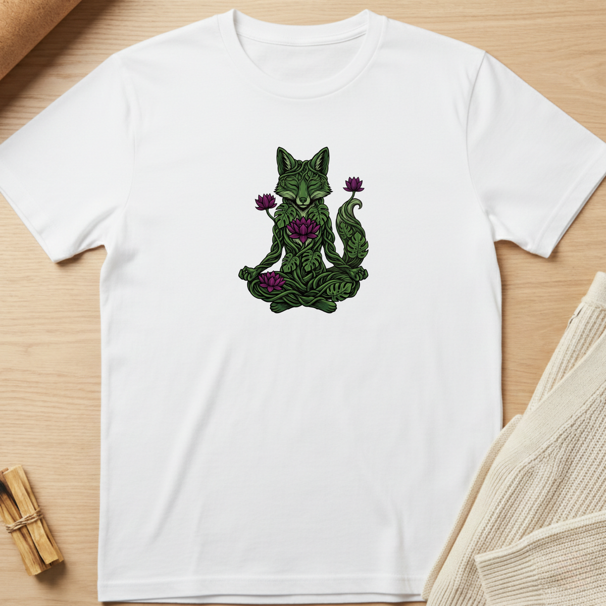 A white short-sleeve crewneck t-shirt laid flat on a light wood surface, featuring a central graphic of a meditating green fox surrounded by purple flowers. Styled flat-lay props include a rolled tan fabric in the top-left and a beige knit sweater in the bottom-right. Minimalist yoga apparel by pemuyo.