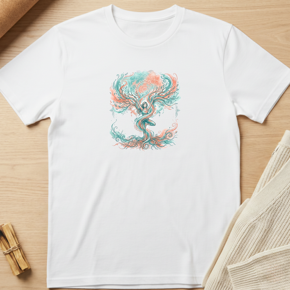 A white short-sleeve crewneck t-shirt laid flat on a light wood surface, featuring a colorful graphic of a meditating figure with flowing teal and coral hair. Styled flat-lay props include a rolled tan paper corner, a small bundle of wooden sticks, and a beige knit fabric on the right. Minimalist yoga apparel by pemuyo.