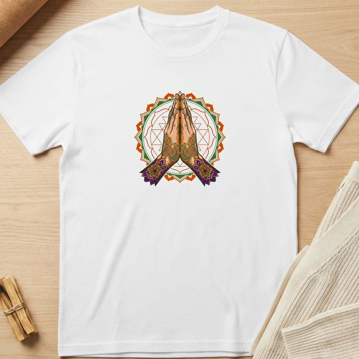 A white short-sleeve crewneck t-shirt laid flat on a light wood surface, featuring a central graphic of praying hands inside a circular mandala design with orange, green, and purple details. Minimalist yoga apparel by pemuyo.