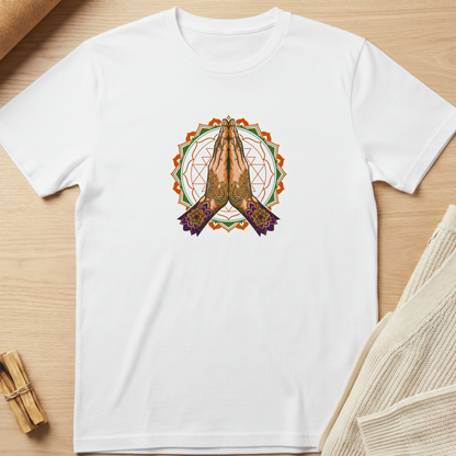 A white short-sleeve crewneck t-shirt laid flat on a light wood surface, featuring a central graphic of praying hands inside a circular mandala design with orange, green, and purple details. Minimalist yoga apparel by pemuyo.